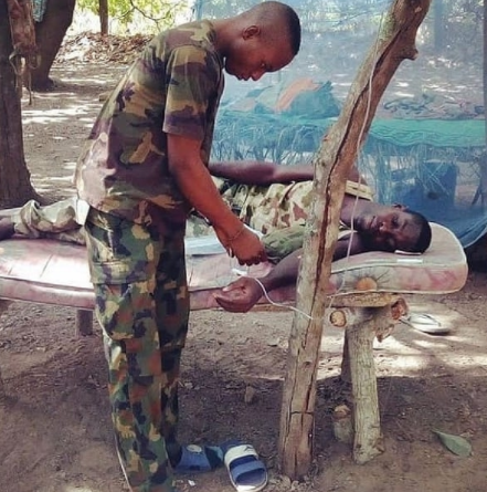 Trending photo of a sick Nigerian soldier receiving treatment on mattress supported by branches while his IV fluid hangs from a tree