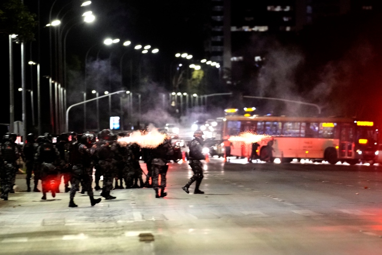 Police and Bolsonaro supporters clash on the street. There is smoke from what are presumably guns being fired to send tear gas canisters in the direction of protesters. The scene looks chaotic.