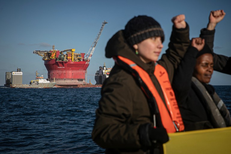 Protesters against oil punch the air. Behind them a platform is being towed by sea to expand an oil field.